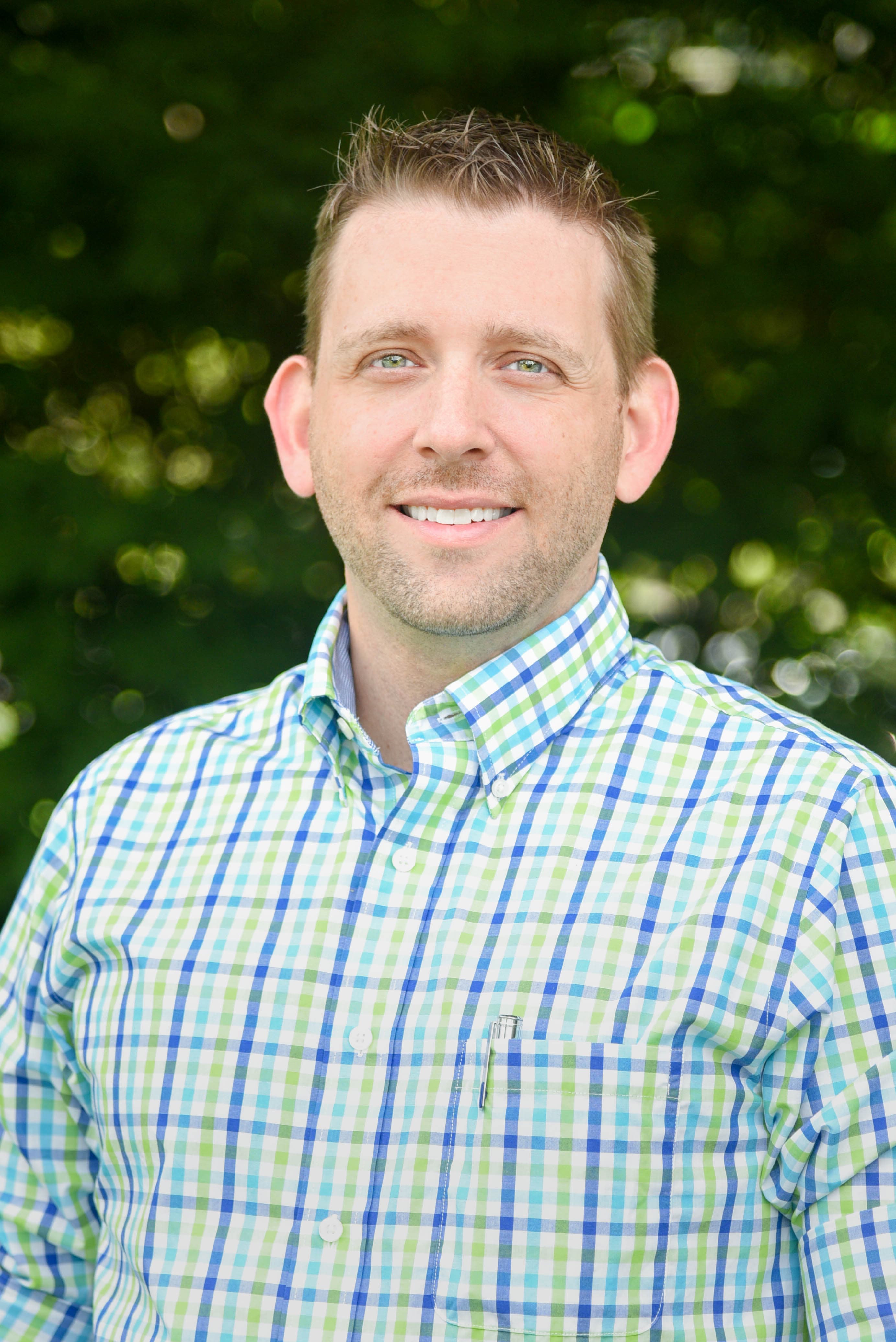 Smiling man with short hair wearing a blue and green plaid shirt against blurred greenery.