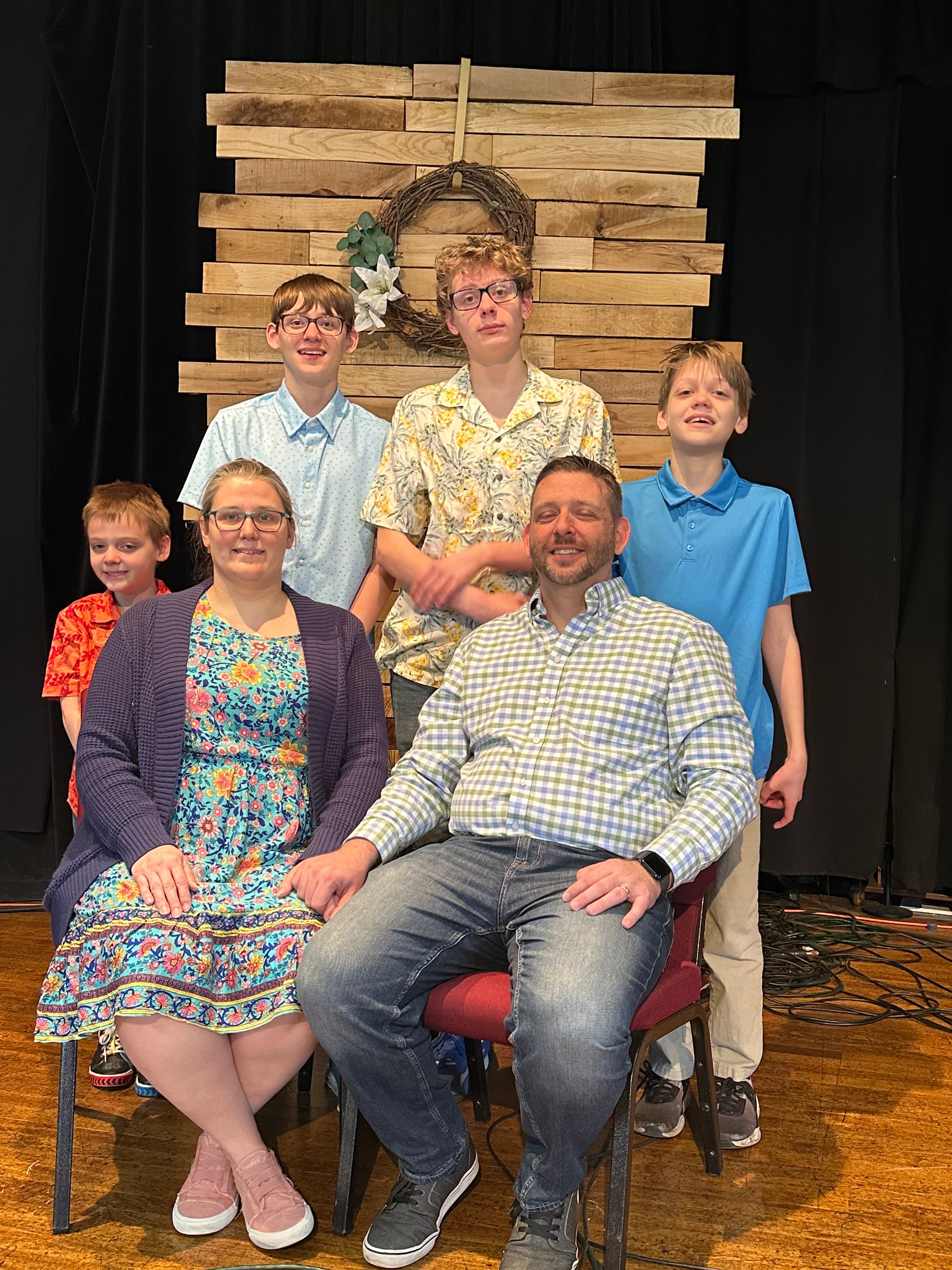 A smiling family of six poses together in front of a rustic wooden plank backdrop.